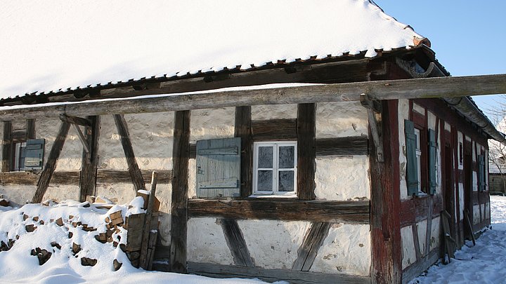 FFM_Schaeferei Fotoaufnahme der Schäferei aus Hambühl am aktuellen Standort. Das Dach, die hölzerne Dachrinne und der Holzstoß vor dem Fachwerk sind schneebedeckt. Auch auf dem Boden ringsum liegt Schnee.