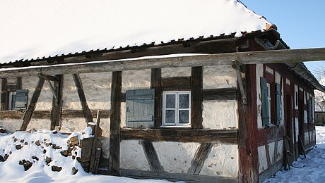 FFM_Schaeferei Fotoaufnahme der Schäferei aus Hambühl am aktuellen Standort. Das Dach, die hölzerne Dachrinne und der Holzstoß vor dem Fachwerk sind schneebedeckt. Auch auf dem Boden ringsum liegt Schnee.