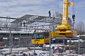 19.3.2018 Fotoaufnahme vom Aufstellen des Schutzdaches. Das große Dach wurde mit Hilfe eines Schwerlastkrans auf den Erdgeschossrohbau gestellt. Das Badhaus ist von einem Gerüst umgeben, auf dem Arbeiter stehen. Rechts befindet sich der Kran, davor liegt schneebedecktes Baumaterial.