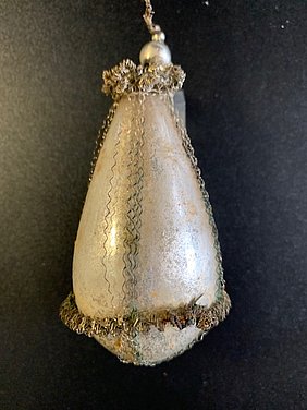 Die Christbaumkugel sieht aus wie ein Tropfen. Sie ist silbern. Sie ist mit glitzerndem Draht umwickelt.