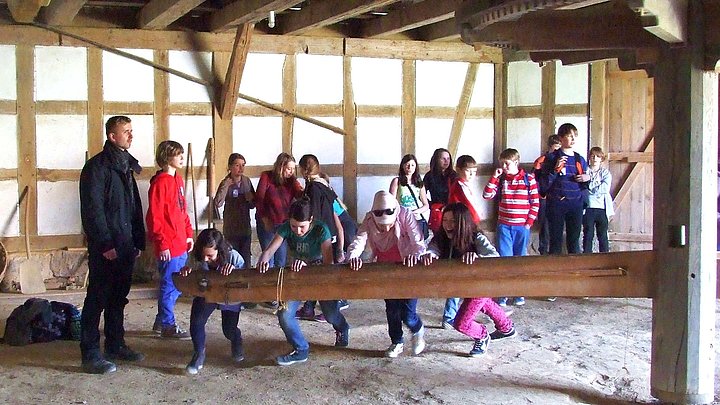 Fotoaufnahme des Handwerker- und Techniktages. In der Mitte des Raums steht ein Göpel. Vier Kinder schieben den großen Balken an, der an einer Antriebswelle aus Holz befestigt ist. Diese wird dadurch in Bewegung gesetzt. An der Fachwerkwand stehen weitere Kinder. 