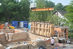 Positionierung der Wanscheiben I Fotoaufnahme von der Positionierung des nordwestlichen Außenwandabschnitts. Mit einem Schwerlastkran wird ein verschaltes Mauerstück auf die Bruchsteinmauer des Erdgeschosses gesetzt. Mehrere Arbeiter am Boden helfen bei der Positionierung.