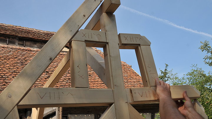 FFM_Zusammensetzen_unseres_Fachwerkmodells Fotoaufnahme des Giebels vom Fachwerkmodell aus Holzbalken. Die linke Seite des Giebels ist schon fertig, die rechte wird gerade zusammengesetzt. In die einzelnen Balken sind Nummerierungen geritzt. Im Hintergrund ist ein mit Wappenbiber-Ziegeln gedecktes Dach erkennbar.
