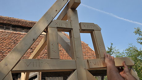 FFM_Zusammensetzen_unseres_Fachwerkmodells Fotoaufnahme des Giebels vom Fachwerkmodell aus Holzbalken. Die linke Seite des Giebels ist schon fertig, die rechte wird gerade zusammengesetzt. In die einzelnen Balken sind Nummerierungen geritzt. Im Hintergrund ist ein mit Wappenbiber-Ziegeln gedecktes Dach erkennbar.