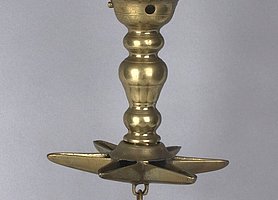 Die Lampe ist aus Messing. Sie hängt an einem Bügel. Sie hat eine Schale, die wie ein Stern aussieht. Sie hat auch eine runde Schale ganz unten. Sie brennt mit Öl.