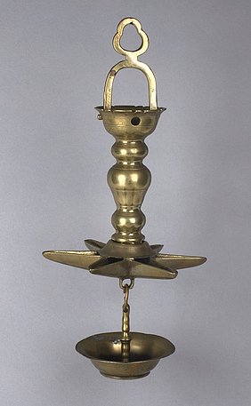 Die Lampe ist aus Messing. Sie hängt an einem Bügel. Sie hat eine Schale, die wie ein Stern aussieht. Sie hat auch eine runde Schale ganz unten. Sie brennt mit Öl.