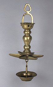 Die Lampe ist aus Messing. Sie hängt an einem Bügel. Sie hat eine Schale, die wie ein Stern aussieht. Sie hat auch eine runde Schale ganz unten. Sie brennt mit Öl.