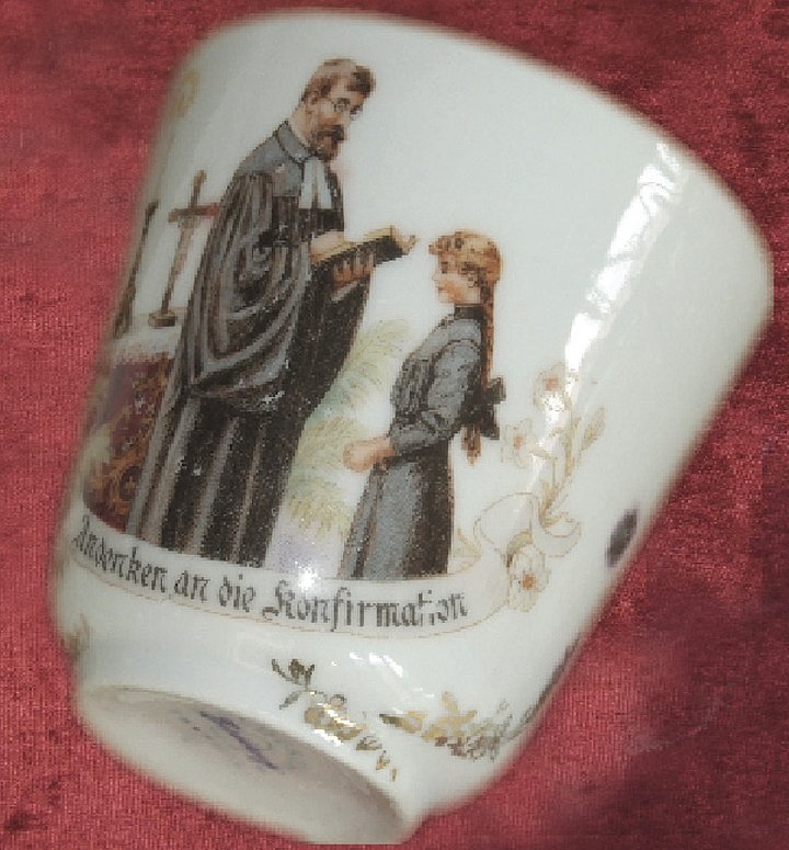 FFM_Vorschau_von_tasse_mit_samt Fotoaufnahme einer Tasse, die auf Samt liegt. Auf der Tasse sind ein Pfarrer und eine Konfirmandin abgebildet. Der Pfarrer im Talar hält eine Bibel in der Hand, aus der er dem Mädchen vorliest. Hinter den beiden ist ein Kreuz erkennbar. Unter dem Bild steht „Andenken an die Konfirmation“