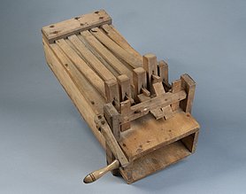 Die Ratsche ist ein Holzkasten. Auf dem Holzkasten sind Holzleisten und eine Kurbel. Die Kurbel zieht an den Holzleisten. Die Holzleisten schlagen auf den Kasten. Es ist ein lautes Knallen zu hören.