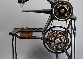 Die Nähmaschine ist schwarz und aus Eisen. Sie wird mit einem Fußpedal angetrieben. Oben ist ein Arm, aus dem Nadel und Faden herauskommen. Darunter ist ein anderer Arm. Auf dem kann das Leder zum Nähen abgelegt werden. Die Maschine hat auch eine kleine Schublade.