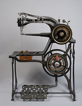 Die Nähmaschine ist schwarz und aus Eisen. Sie wird mit einem Fußpedal angetrieben. Oben ist ein Arm, aus dem Nadel und Faden herauskommen. Darunter ist ein anderer Arm. Auf dem kann das Leder zum Nähen abgelegt werden. Die Maschine hat auch eine kleine Schublade.