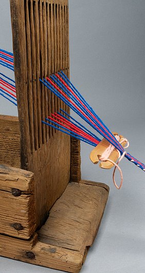 Durch den Kamm laufen die Fäden. Es gibt oben eine Reihe Fäden, und es gibt unten eine Reihe Fäden. Sie werden mit einem weiteren Faden zusammengeführt. Am Ende entsteht ein Band.