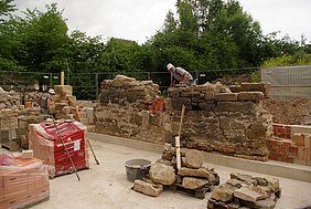 Die beiden Wandteile nach dem Entfernen der Verschalung Fotoaufnahme zweier Wandteile nach dem Entfernen der Verschalung. Arbeiter mauern die Fehlstelle zwischen beiden Bauteilen mit passenden Bruchsteinen aus. In der Grube befinden sich verpackte Hochlochziegel und zwei Paletten mit Sandsteinquadern. Im Hintergrund stehen Bäume.