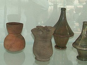 Vierpassbecher im Vordergrund, dahinter ein tönerner Beutelbecher und 2 konische Glasflaschen