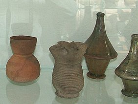 Vierpassbecher im Vordergrund, dahinter ein tönerner Beutelbecher und 2 konische Glasflaschen
