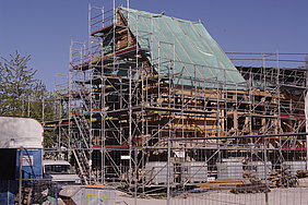 Das auf allen Seiten eingerüstete Badhaus aus Wendelstein, ein Drittel des Dachwerks ist schon aufgestellt und mit einer Plastikplane bedeckt.