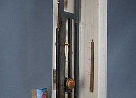 Der Apparat ist aus Holz. An ihm ist eine Scheibe mit Nägeln. Auf die Nägel werden Pinselstiele gesteckt. Der Apparat taucht die Stiele in Lack.