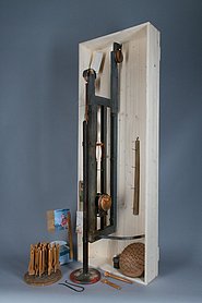 Der Apparat ist aus Holz. An ihm ist eine Scheibe mit Nägeln. Auf die Nägel werden Pinselstiele gesteckt. Der Apparat taucht die Stiele in Lack.
