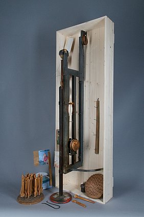 Der Apparat ist aus Holz. An ihm ist eine Scheibe mit Nägeln. Auf die Nägel werden Pinselstiele gesteckt. Der Apparat taucht die Stiele in Lack.