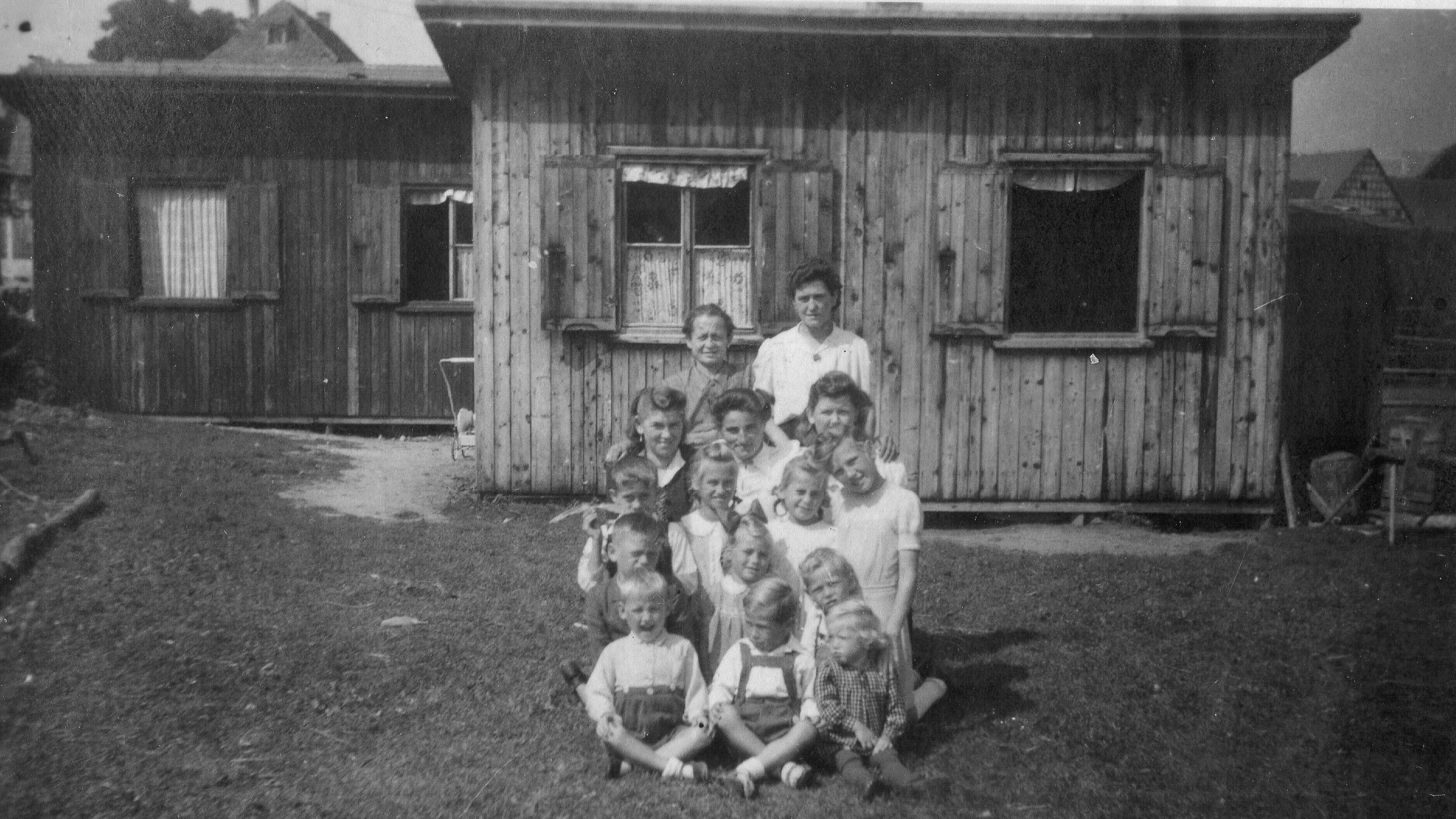 Die beiden Behelfsheime sind kleine Holzhäuser. Davor stehen Frauen und Kinder. Sie wohnen in den Häusern.