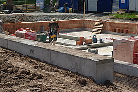 Füllung der Baugrube mit Sand und Vormauerung mit Hochlochziegel Fotoaufnahme der Füllung der Baugrube mit Sand und Vormauerung mit Hochlochziegel. In der Grube stehen ein Mann mit kurzen Arbeitshosen und Strohhut sowie verpackte Steine und andere Utensilien. Vor dem Bauzaun im Hintergrund stapelt sich weiteres Baumaterial.