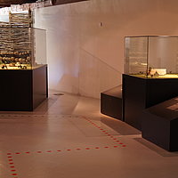 FFM_Archaeologisches_Dorf_Virnsberg_Ausstellung Fotoaufnahme der Ausstellung des Archäologiemuseums, welches sich in der Schafscheune aus Virnsberg befindet. Gezeigt werden zwei Vitrinen, in denen sich Modele von Siedlungen bzw. Häusern befinden. Auf dem Boden ist ein Leitsystem durch die Ausstellung erkennbar.