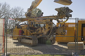 Foto des Rammgeräts zum Einbringen der Stahlrohrpfähle Fotoaufnahme eines Rammgeräts zum Einbringen der Stahlrohrpfähle. Im Vordergrund steht ein Bauzaun. Dahinter befinden sich links das Rammgerät und rechts ein Container der Firma Max Bögl. Links im Hintergrund sind kahle Bäume erkennbar.