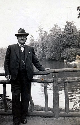 Schwarz-Weiß Fotografie von einem Mann im Anzug mit Hut, an einem Geländer lehnend, vor einem Gewässer. 