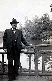 Schwarz-Weiß Fotografie von einem Mann im Anzug mit Hut, an einem Geländer lehnend, vor einem Gewässer. 