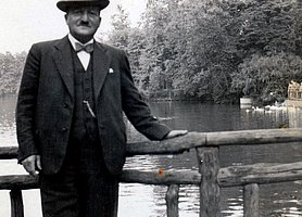 Schwarz-Weiß Fotografie von einem Mann im Anzug mit Hut, an einem Geländer lehnend, vor einem Gewässer.