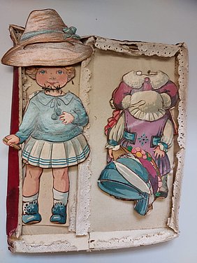 In der Schachtel liegt eine Puppe. Sie ist auf Pappe gedruckt. Daneben liegen viele Kleidungsstücke. Sie sind auch auf Pappe gedruckt. Die Kleidungsstücke sind bunt. Alles ist flach und kann aufeinander gelegt werden.
