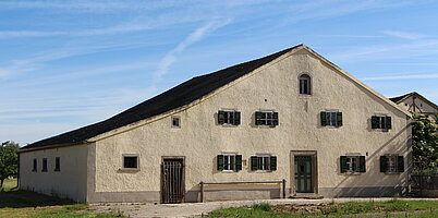 FFM_Bauernhaus aus Reichersdorf Fotoaufnahme des Bauernhauses aus Reichersdorf am aktuellen Standort. Das breitgelagerte eingeschossige Jurahaus hat zwei Eingänge an der Giebelseite. Links ist die Stalltür, rechts die Haustür aus Holz. Die Fenster auf der Wohnseite haben geöffnete Läden.