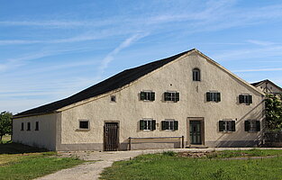 Fotoaufnahme des Bauernhauses aus Reichersdorf am aktuellen Standort. Das breitgelagerte eingeschossige Jurahaus hat zwei Eingänge an der Giebelseite. Links ist die Stalltür, rechts die Haustür aus Holz. Die Fenster auf der Wohnseite haben geöffnete Läden. 