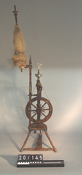 Das Spinnrad ist aus Holz. Es ist verziert. Manche Teile sind aus Zinn.