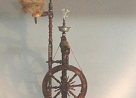 Das Spinnrad ist aus Holz. Es ist verziert. Manche Teile sind aus Zinn.