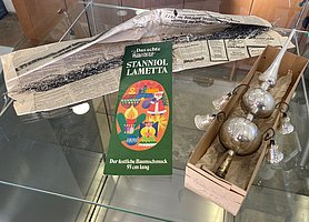 Das Lametta besteht aus silbernen, glänzenden Streifen. Davor liegt eine Schachtel. In ihr ist eine silberne Christbaumspitze. Sie besteht aus einer Spitze und zwei Kugeln. An den Kugeln sind vier Glöckchen.