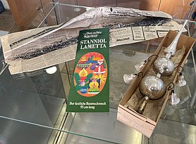 Das Lametta besteht aus silbernen, glänzenden Streifen. Davor liegt eine Schachtel. In ihr ist eine silberne Christbaumspitze. Sie besteht aus einer Spitze und zwei Kugeln. An den Kugeln sind vier Glöckchen.