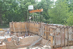 Positionierung der Wanscheiben II Fotoaufnahme der Positionierung der Wandscheiben. Die Hälfte der Wände des Erdgeschosses sind bereits an ihrem Platz. In eine Fehlstelle wird gerade ein weiteres verschaltes Bauteil auf das Bruchsteinmauerwerk gesetzt. Die stehenden Wände werden von Leitern und Stützpfosten gehalten.