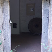 FFM_West_Oelmuehle_Muehlraum_Eingang Fotoaufnahme des hinteren Eingangs zum Mühlraum der Ölmühle von der Fledrichsmühle am aktuellen Standort. Links und rechts neben der Tür wächst u.a. Klee und Gras. Im Inneren des Raums lehnt ein Mühlstein an der Wand, darüber wird an einer Tafel der Mühlgang erklärt.