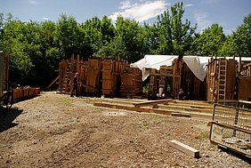 Bauteillager Fotoaufnahme des Bauteillagers für das Badhaus aus Wendelstein. Die massiven Wandscheiben mit Verschalung sind nebeneinander aufgereiht. Über dem rechten Teil liegt eine Plane. Im Hintergrund ist eine Baumreihe erkennbar.