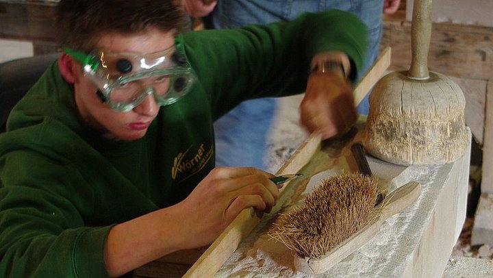 Fotoaufnahme der Steinwerkstatt. Ein Jugendlicher mit Schutzbrille kniet vor einem Steinblock und misst ihn aus. Auf dem Steinblock liegen ein Hand-Kehrer, ein Meißel und ein Knüpfel. Im Hintergrund arbeitet ein weiterer Mann.