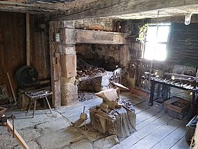 Die Schmiede ist eine Werkstatt. In der Mitte steht ein Amboss. Dahinter ist eine Esse. In der Esse wird Eisen heiß gemacht. An den Wänden sind Regal. In den Regalen hängen Werkzeuge. Das Sind Hämmer und Zangen. Der Boden ist gepflastert.
