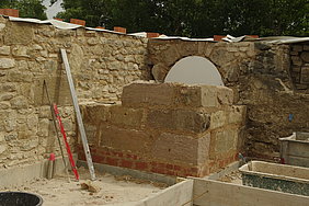 Fotoaufnahme des wiederaufgebauten Ofens mit seiner äußeren Schale aus Sandsteinquadern. Der Ofen befindet sich in einer Ecke. Der untere Teil wurde mit Ziegelsteinen gemauert, darüber sind drei Reihen Sandstein erkennbar. Links daneben stehen zwei Wasserwagen und eine Thermoskanne.