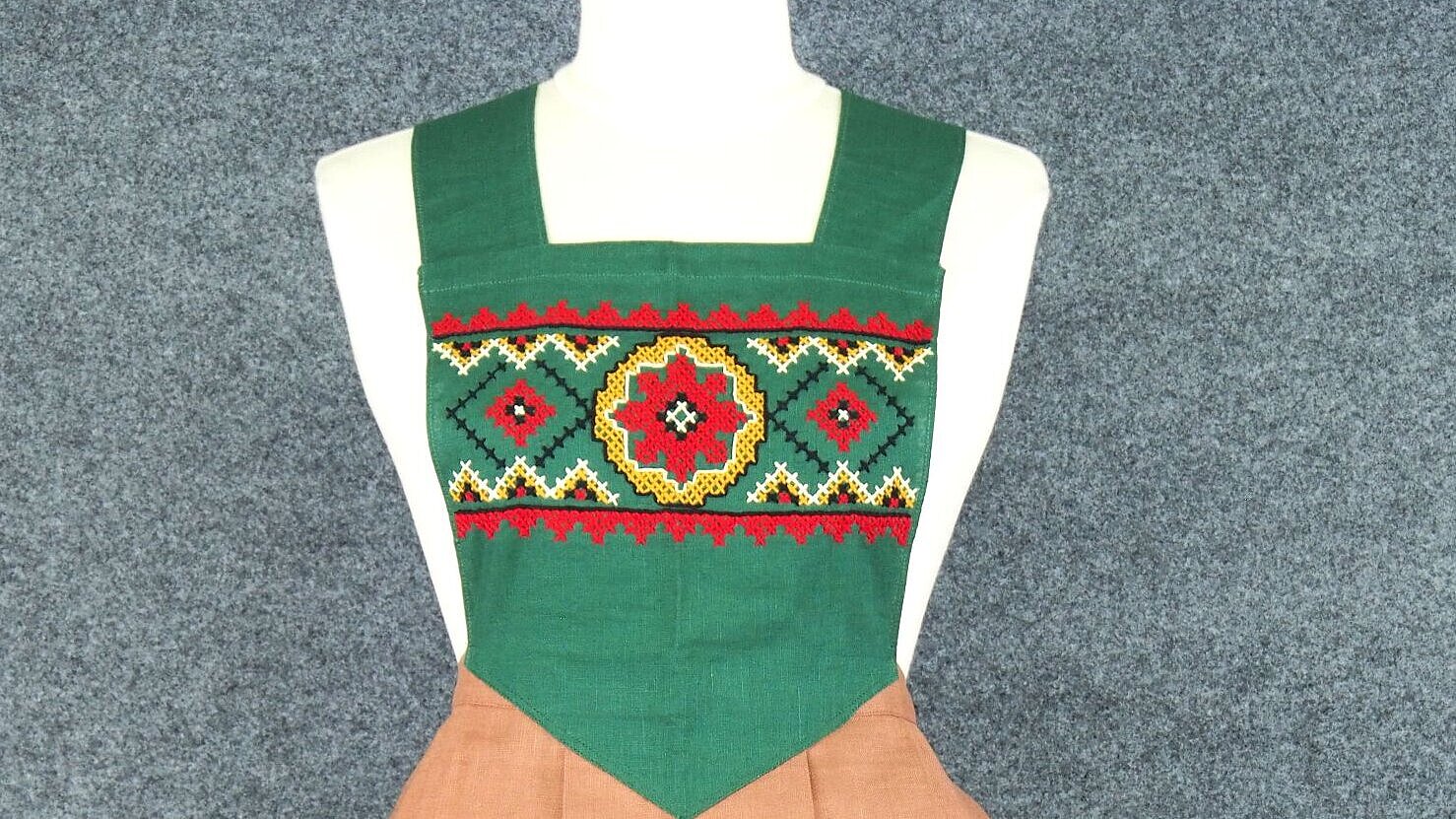 Die Schürze ist grün, rot und braun. Sie hat zwei Taschen und ein Blumenmuster.