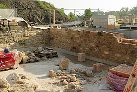 Blickrichtung Nordost Fotoaufnahme der unteren Wandabschnitte der zukünftigen Badstube. Um das Eck der Baugrube führt eine Steinmauer. In der Grube liegen Sand- und Ziegelsteine auf Paletten und am Boden. Außerhalb der Grube sind Teile des Bauzaun und des Erdhügels erkennbar.