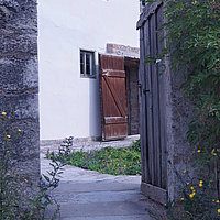 FFM_West_Kleinrindferfeld_Tor Fotoaufnahme des Hofzugangs zum Bauernhaus aus Kleinrinderfeld am aktuellen Standort. Die in eine Mauer eingelassene Holztür ist nach innen geöffnet. Vor der Schwelle liegt eine Rampe aus Holz, links und rechts davon blühen verschiedenen Wildblumen. Die Stalltür im Inneren ist ebenfalls offen.