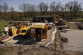 Fotoaufnahme der Baustelleinrichtung für die Pfahlgründung durch die Firma Max Bögl. Links neben der Baugrube stehen verschiedene Maschinen, Fahrzeuge und Werkzeuge der Firma. Die meisten wurden mit dem Firmennamen beschriftet. Im Hintergrund sind kahle Bäume und ein Strommast. 
