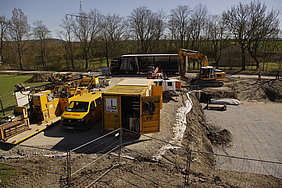 Foto der Baustelleneinrichtung Fotoaufnahme der Baustelleinrichtung für die Pfahlgründung durch die Firma Max Bögl. Links neben der Baugrube stehen verschiedene Maschinen, Fahrzeuge und Werkzeuge der Firma. Die meisten wurden mit dem Firmennamen beschriftet. Im Hintergrund sind kahle Bäume und ein Strommast.