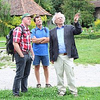 FFM_Fuehrungen Fotoaufnahme zweier Besucher mit einem Museumsführer. Die drei Männer betrachten etwas außerhalb des Bildes. Einer steht auf dem Kiesweg, die anderen beiden im Gras. Im Hintergrund befinden sich eine Bierzeltgarnitur sowie Büsche, Blumen und Gebäude.