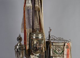 Die Sachen gehörten einem Rauchclub. In dem Club hat man gemeinsam Tabak geraucht. In der Mitte steht eine sehr große Pfeife. An ihr hängen viele Fahnen. Daneben steht ein Pokal. Es gibt noch eine kleine Fahne. Außerdem gibt es noch zwei kleine Pfeifen.